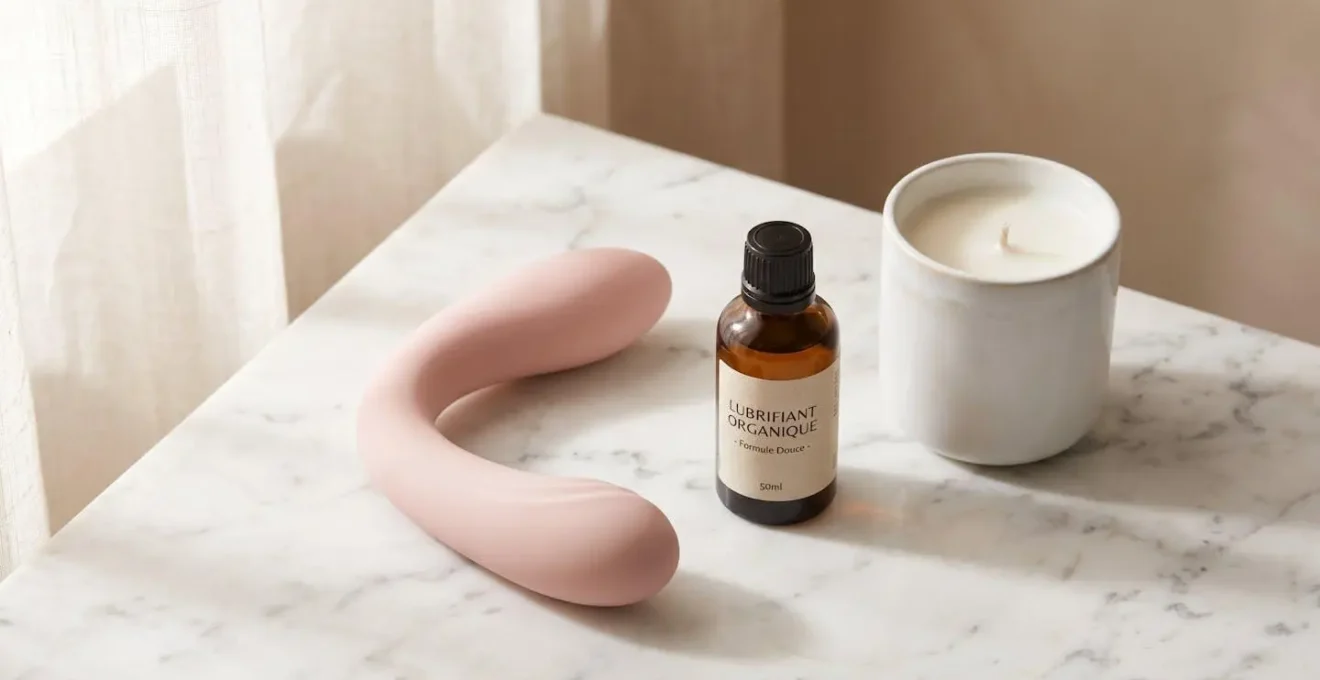 Nature morte produits érotiques premium sur marbre blanc : vibromasseur design rose poudré en silicone médical, flacon de lubrifiant biologique en verre ambré, bougie de massage céramique, éclairage doux naturel